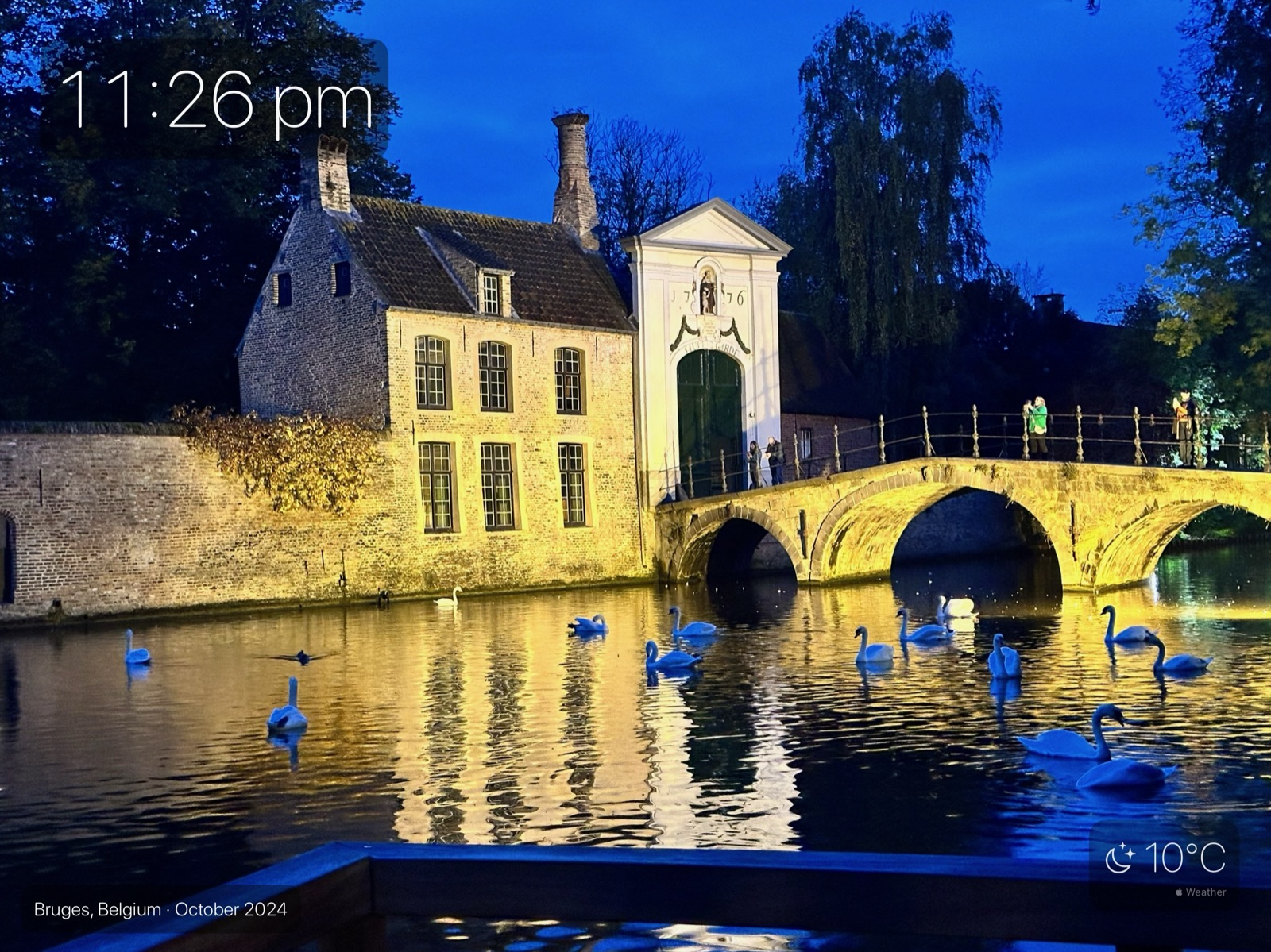 An iPad showing a night photograph of swans on a canal in Bruges, with the time in the top corner and the temperature in the bottom corner.